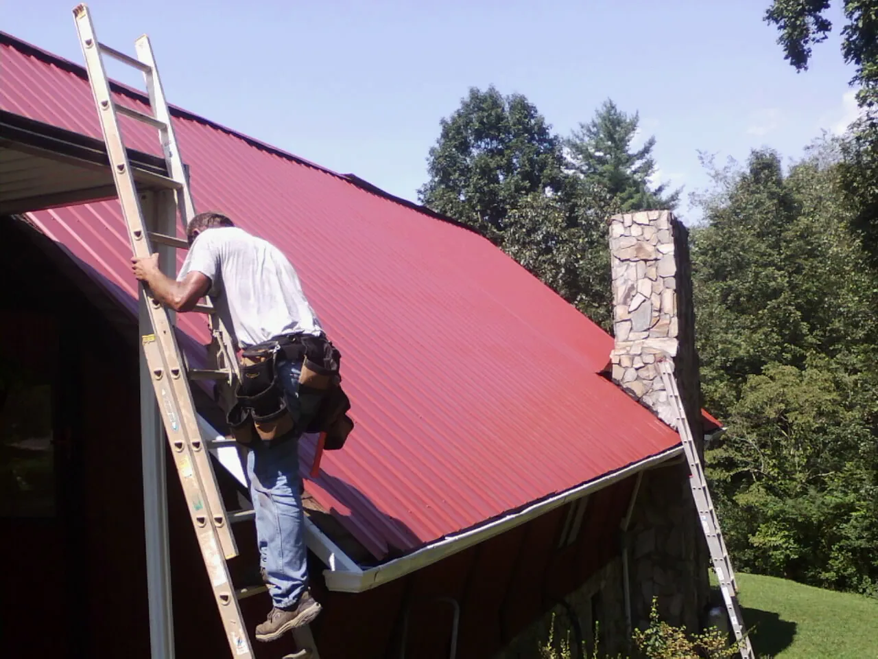 Roofer on a ladder beside a red metal roof and stone chimney — Soffit & Fascia Repair by Roofing Brothers in Leetonia